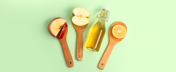Bottle of apple cider vinegar and spoons with fruits on green background, top view