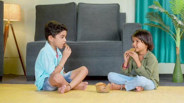 Young sibling kids having snacks or biscuit while sitting on floor at home by looking around after school - concepts of healthy eating togetherness and childhood lifestyle.