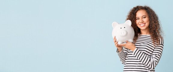 Happy young African-American woman with big piggy bank on light blue background with space for text