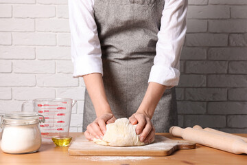 Woman kneading dough at wooden table near white brick wall, closeup