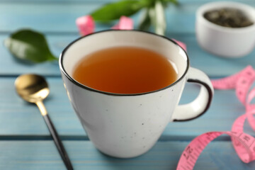 Cup of herbal diet tea and measuring tape on light blue wooden table, closeup. Weight loss concept