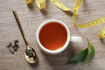 Cup of herbal diet tea and measuring tape on wooden table, flat lay. Weight loss concept