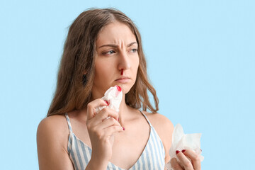 Young woman with nosebleed and tissue on blue background, closeup