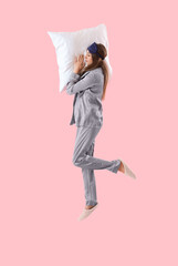 Young woman in pajamas with pillow jumping on pink background