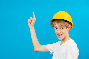 Happy handsome teen builder in a yellow helmet is looking at the camera and points fingers up, standing in a studio on a blue background