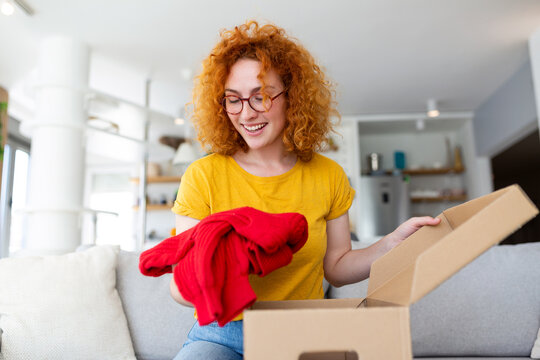 Beautiful young cheerful red hair woman unboxing a package in the living room of a new sweater.