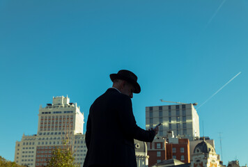 Silhouette of adult man in suit and hat looking at mobile against buildings and blue sky. Madrid, Spain