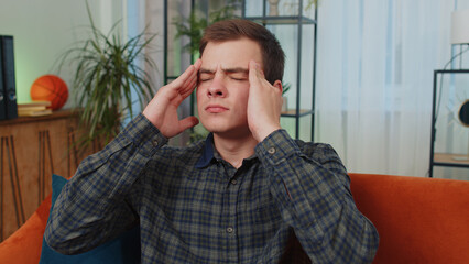 Displeased caucasian teenager man rubbing temples to cure headache problem, suffering from tension and migraine, stress, grimacing in pain, high blood pressure. Young guy at home living room on couch