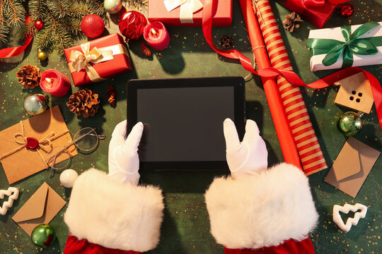 Santa Claus holding tablet computer on green table with Christmas decor and gifts, top view