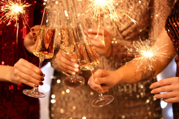 Young women in elegant dresses with glasses of champagne and burning Christmas sparklers, closeup