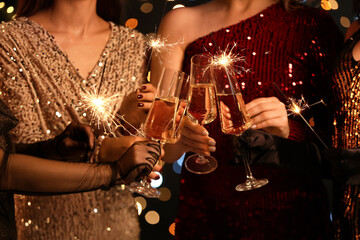 Young women in elegant dresses with burning Christmas sparklers and glasses of champagne, closeup