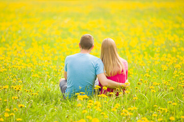 Fototapeta premium Young adult couple hugging each other and sitting at meadow of fresh yellow blooming dandelions in sunny spring day. Looking far away. Peaceful atmosphere in nature. Back view. Romantic lovely moment.