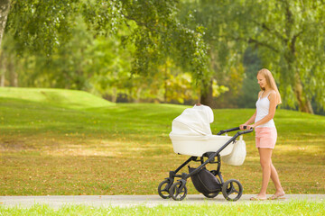 Fototapeta na wymiar Smiling young mother pushing white baby stroller and walking on sidewalk at beautiful town park. Warm sunny summer day. Spending time with newborn and breathing fresh air. Enjoying stroll. Side view.