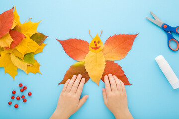 Child hands creating butterfly shape from colorful leaves on light blue table background. Pastel...