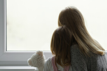 Little girl holding white teddy bear. Young adult mother and daughter together looking out from window at home. Back view. Waiting, longing or sadness concept. Closeup.