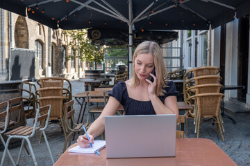 Portrait of happy young office assistant working remotely online sitting with laptop and coffee outside on terrace in a european city making notes in agenda in same time speaking with smile on phone