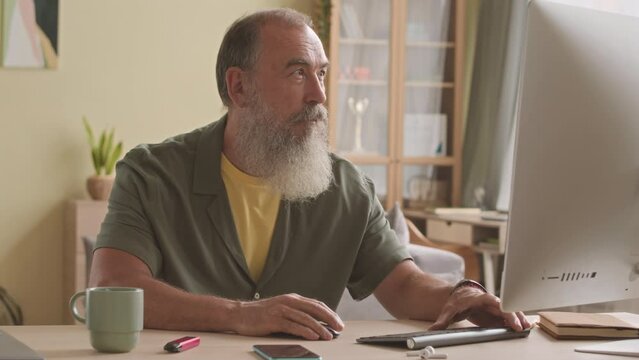 Modern boomer man with long grey beard using computer and checking notifications on smartphone, sitting at desk in living room