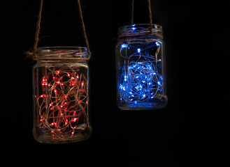 Two glass jars with a luminous garlands inside with many lights hanging on a jute rope against a...