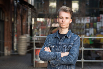 Portrait of a young blond guy with a stylish hairstyle with crossed arms in a denim jacket.