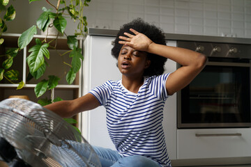 Sweaty overheated young African woman using electric fan ventilator, black girl suffering without...