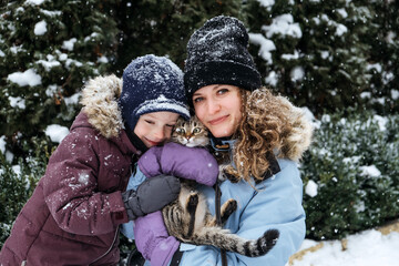 Obraz premium Portrait of happy family mother, son and cat in snowy winter park. Outdoors Portrait of mom and kid boy with cat hugging in winter snow nature background
