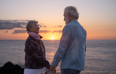 Fototapeta premium Lovely senior couple standing on the rocks at sea hand in hand enjoying nature and freedom at sunset light. Relaxed lifestyle for a caucasian couple of retirees