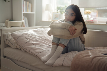 Pensive caucasian teenage girl sitting on bed and looking away