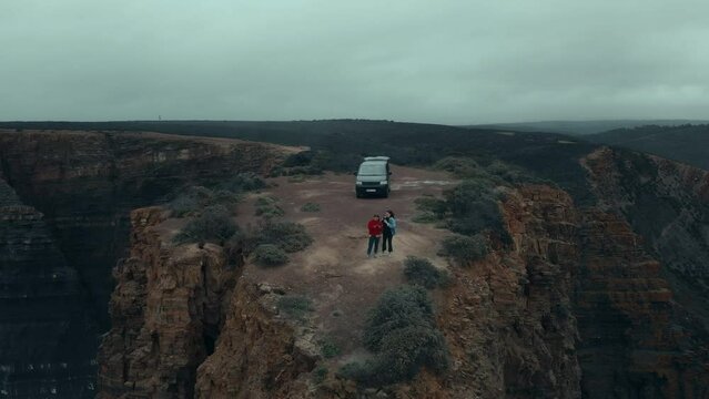 Aerial drone shot of couple stand on top of adventure van at edge of cliff on epic and dramatic coastline. Cinematic shot of moody travel destination. Vanlife lifestyle for millennials, happy friends