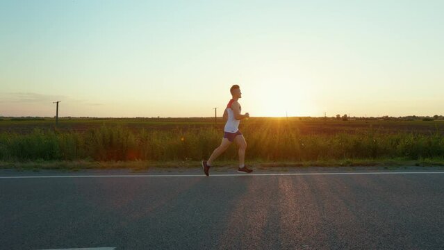 Side view of strong man running, taking part in marathon in countryside. Male athlete wearing sportswear jogging, working out, training at sunset. Concept of sport and running.