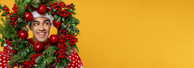 Close up merry fun young man wearing red knitted sweater Santa hat posing hold look aside through Christmas wreath isolated on plain yellow background Happy New Year 2023 celebration holiday concept.