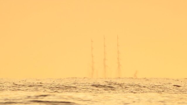 Mirage of sailboat masts in the sea beyond the horizon. Ship sailing in the sea. Small waves on the golden surface of warm water with bokeh lights from the sun. Sea, nature and outdoor travel.