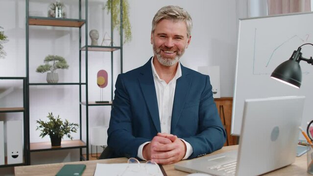 Mature business man working on laptop computer smiling friendly at camera and waving hands gesturing hello, hi, greeting or goodbye, welcoming with hospitable expression at home office workplace desk