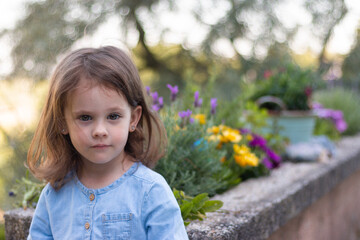 A little beautiful preschooler girl stands in a denim blue dress against the background of flowers