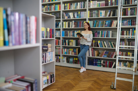 Young librarian standing near the bookshelves with a gadget in hands