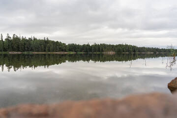 Beautiful view of the quiet lake reflecting the gray sky on a cloudy day outside. The rocky shore in the foreground is out of focus. Natural background.