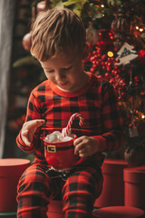 Portrait candid happy child in red plaid pajama hold Xmas mug with marshmallows and candy cane