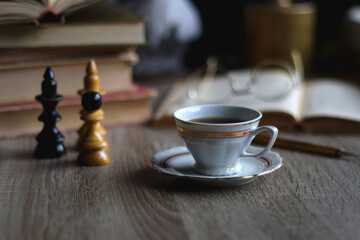 Stack of vintage books, cup of tea or coffee, lit candles, reading glasses and chess pieces on wooden table. Dark academia concept. Selective focus.