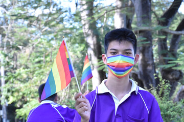 Portrait of young asian boy wears rainbow mask and holding rainbow flag, LGBT symbol, in the park, soft and selective focus, concept for LGBT community celebration and respecting gender diversity.