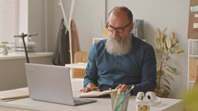 Medium shot of old adult Caucasian businessman with long gray beard using laptop at workplace