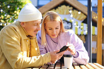 A young man and his girlfriend are sitting at the wooden table of a street cafe in and looking at the phone screen together