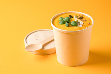 Creamy pumpkin fall soup in craft paper food container with bamboo spoon against orange background...