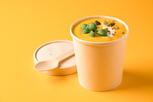 Creamy pumpkin fall soup in craft paper food container with bamboo spoon against orange background with copy space. Butternut squash cream soup. Food delivery, takeaway soup. Mockup image