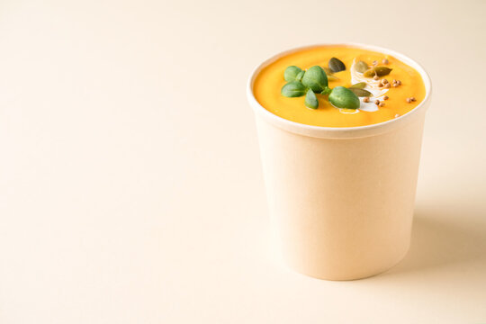 Creamy pumpkin fall soup in craft paper food container against light background with copy space. Butternut squash cream soup garnished with seasonings. Food delivery and soup to go. Mockup