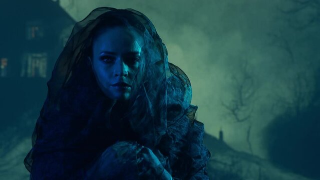 Beautiful witch making the witchcraft over the smoky background. Scary house on the hill. Halloween image.