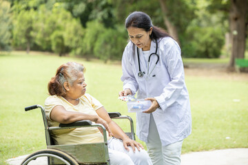 Indian doctor or nurse give medicine to senior female patient in a wheelchair outdoor at park.