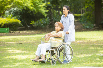 Indian caregiver nurse taking care of senior female patient in a wheelchair outdoor at park, Asian doctor help and support elderly mature older people. rehabilitation and health care.