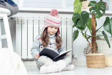 Interested, serious, cute little girl in woolen hat and warm clothes with snowman reading book next...