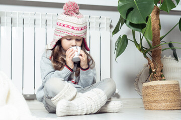 Lovely little girl in wool hat, socks and pullover drink hot tea, sit with crossed legs near heating convector, radiator