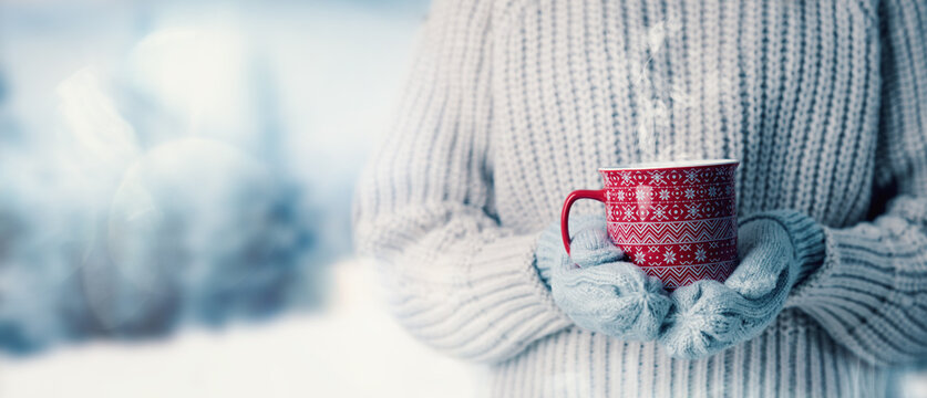 woman in wool sweater and mitten gloves holding a cup of hot steaming drink on snowy winter landscape background. banner with copy space
