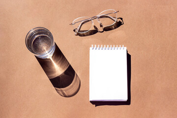 Empty notepad, glass of water and eyeglasses with strong shadows on brown background. Creative mockup. Top view, flat lay, Still life